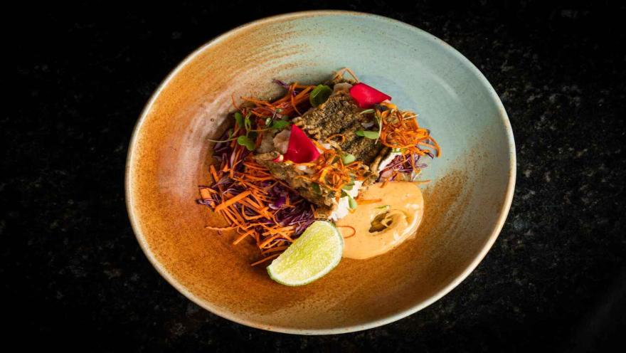 Plato de cocina contemporánea con pescado, vegetales frescos, flores comestibles, salsa cremosa y una rodaja de limón.