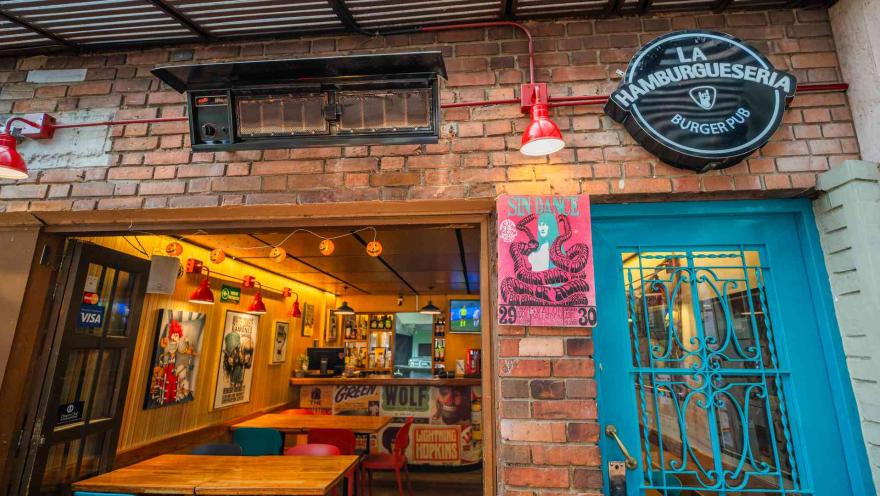 Entrada con puerta metálica azul, letrero luminoso de La Hamburguesería Burger Pub y vista al comedor iluminado.
