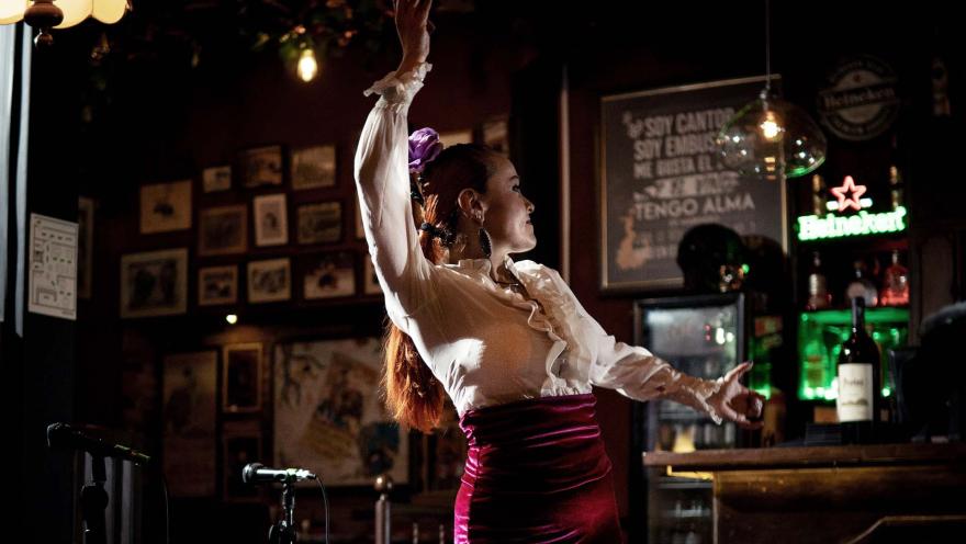 Mujer bailando flamenco en el interior de un bar, rodeada de luces cálidas, botellas y decoración tradicional.