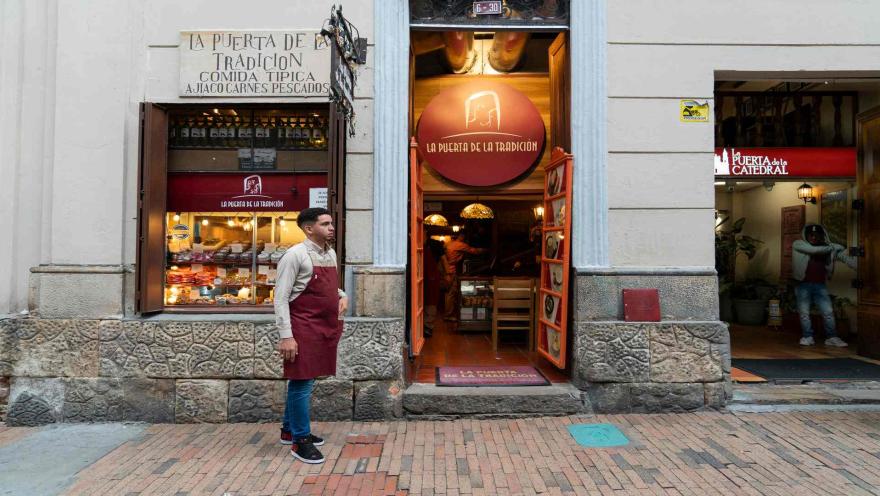 Fachada del restaurante La Puerta de la Tradición con un mesero de delantal rojo parado en la entrada. 