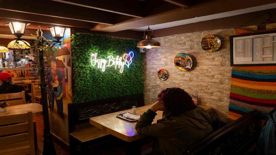 Mujer sentada a la mesa en un comedor con pared verde, letrero de neón Happy Birthday y platos decorativos. 