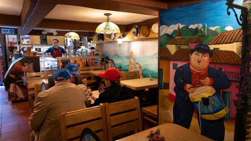 Clientes sentados en mesas de madera junto a un mural pintado de un personaje tradicional con un tambor. 