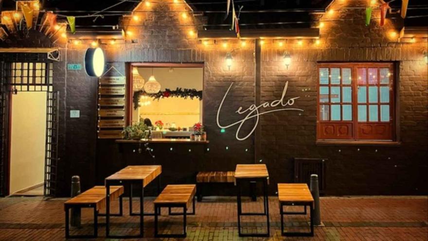 Fachada nocturna de ladrillo con luces cálidas, letrero luminoso de Legado y mesas en la terraza exterior. 