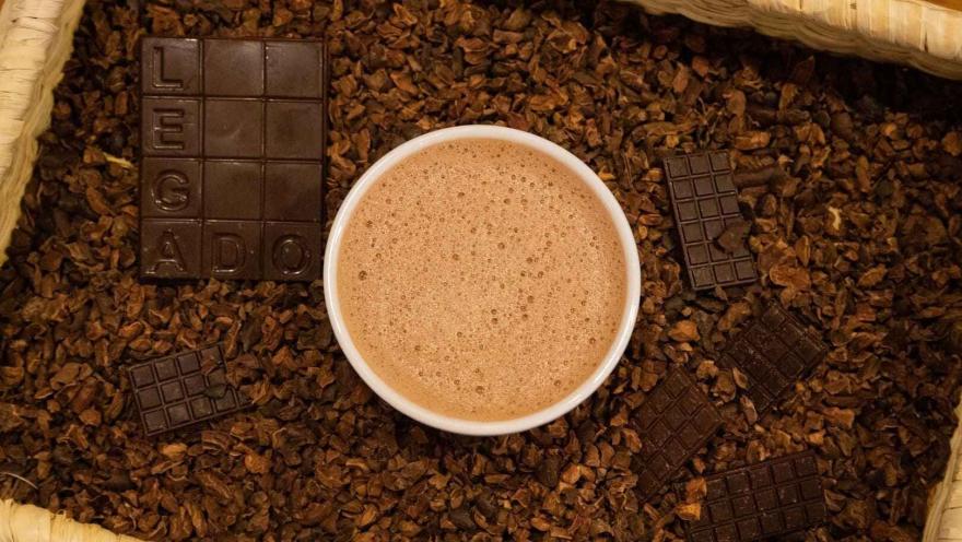 Vista superior de una taza de chocolate caliente rodeada de barras de chocolate y nibs de cacao en una cesta. 