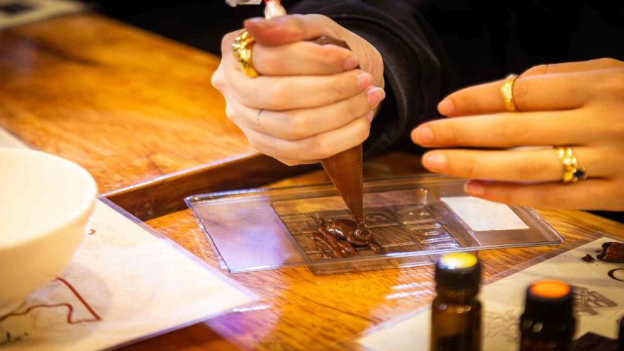 Manos vertiendo chocolate derretido con una manga pastelera en un molde para crear una barra artesanal. 