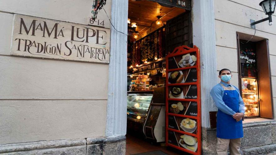 Plano medio de la entrada con placa de Mamá Lupe Tradición Santafereña, menú en marco rojo y empleado junto al portal. 