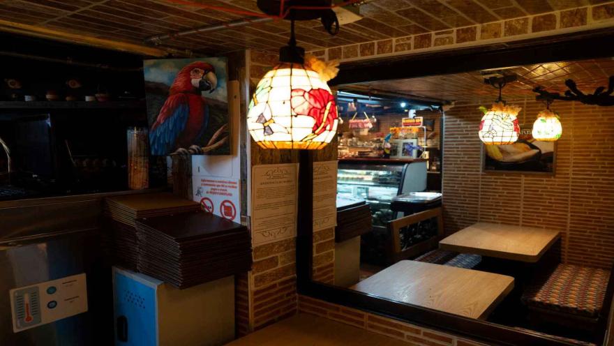 Rincón del restaurante con mesa de madera, pared de ladrillo, pintura de guacamaya y lámpara de vitral colgante. 