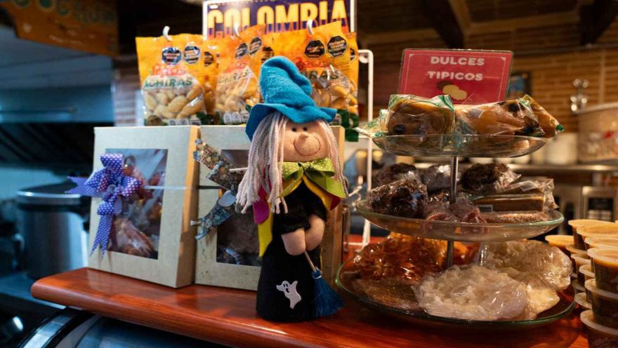  Primer plano de una muñeca de bruja artesanal sobre mostrador con paquetes de achiras y exhibidor de dulces. 