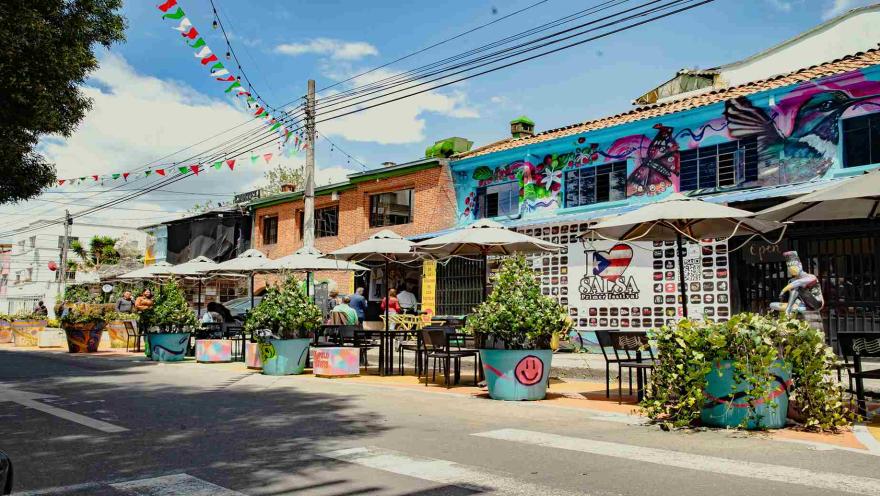 Calle colorida con sombrillas de terraza, macetas pintadas con caras felices y un mural que dice "SALSA".