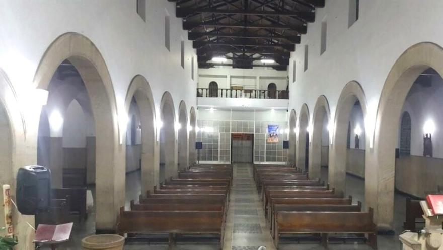 Plano general interior de la iglesia vista desde el altar, en el que se observan varias columnas en forma de arco y dos filas de sillas en madera. 