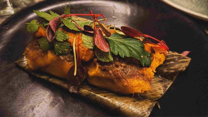 A dish with rich, bold flavors, served on a leaf and accompanied by roasted vegetables, fresh herbs, and delicate microgreens.