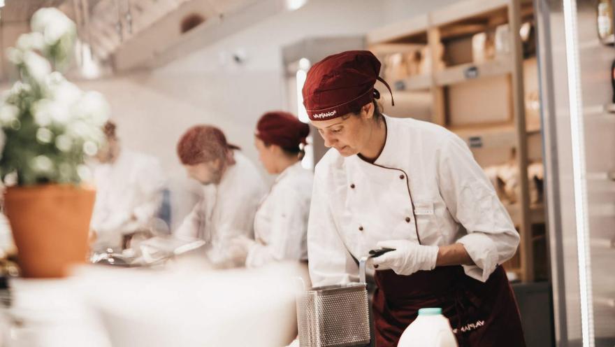 Chef de Vapiano preparando alimentos en la cocina, con otros integrantes del equipo trabajando al fondo.