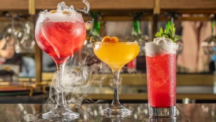 Selección de cócteles de colores vibrantes, servidos en diferentes copas y vasos, con decoraciones frescas y efectos de humo.
