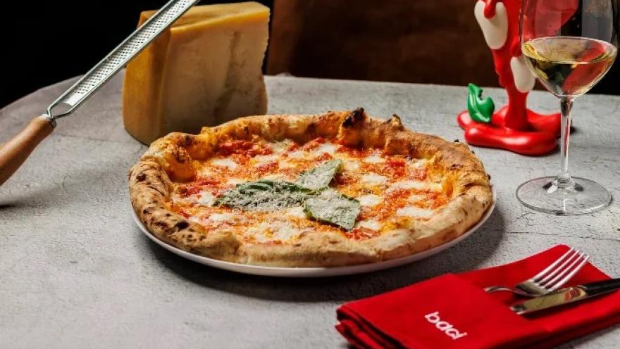 Pizza napolitana de masa artesanal, cubierta con tomate, mozzarella y albahaca, acompañada de queso parmesano y una copa de vino.