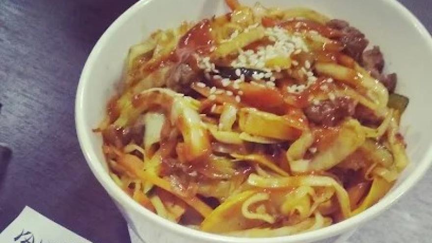 Plato de fideos salteados con carne, vegetales y salsa, acompañado de ajonjolí, servido en un tazón blanco.