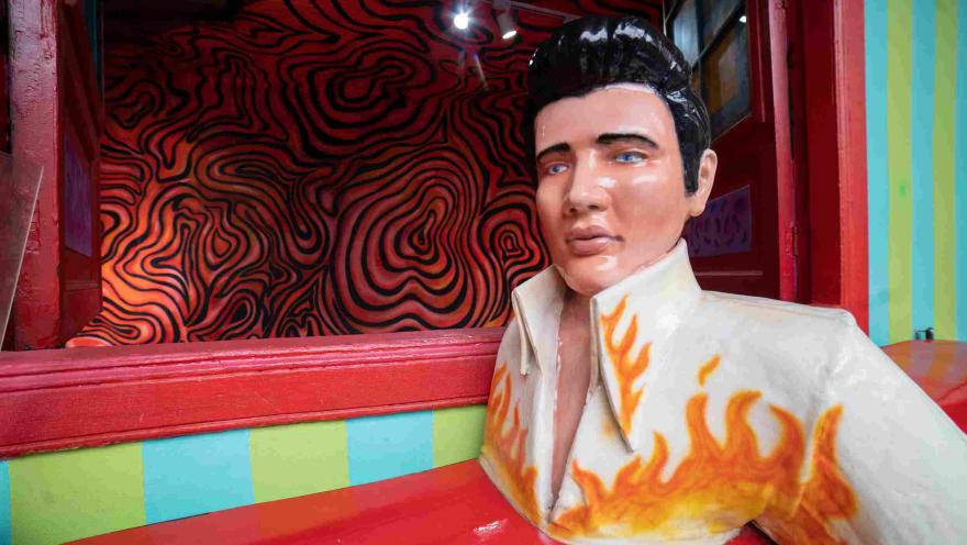 Escultura de una figura masculina con apariencia de estrella de rock, vestida con una chaqueta blanca decorada con llamas naranjas, ubicada en uno de los coloridos escenarios del Museo de la Selfie en Bogotá. Detrás se observa una pared con diseños psicodélicos en tonos rojos y negros.