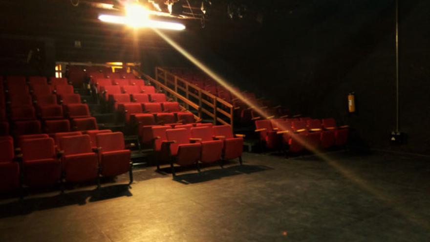 Sala de teatro con filas de sillas rojas, iluminación escénica y espacio dispuesto para disfrutar de una función.