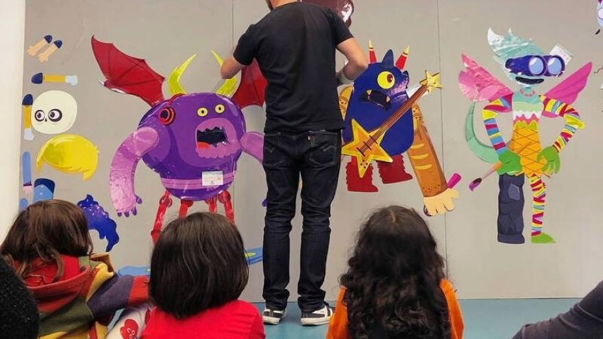 Un grupo de niñas y niños observa a un artista mientras crea una colorida obra con personajes fantásticos y monstruos de diferentes formas, tamaños y colores, en un espacio dedicado al arte y la creatividad infantil.