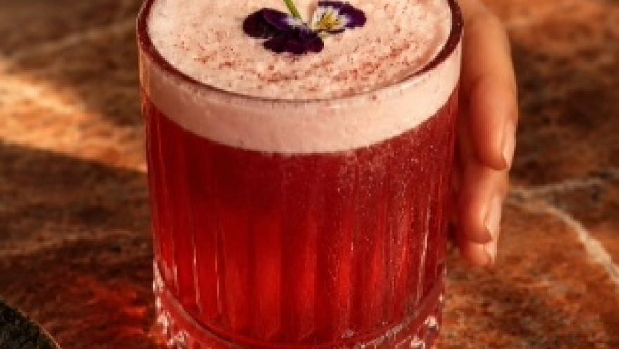 Cóctel de color rojo intenso, servido en vaso bajo y decorado con espuma y una flor comestible.