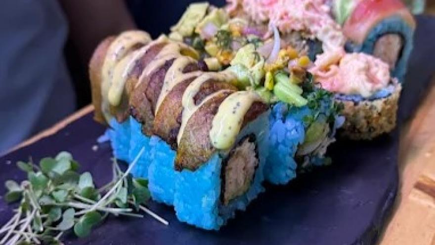 Sushi roll de arroz azul con relleno de pescado y vegetales, cubierto con salsas, semillas y toppings, servido sobre una bandeja negra.