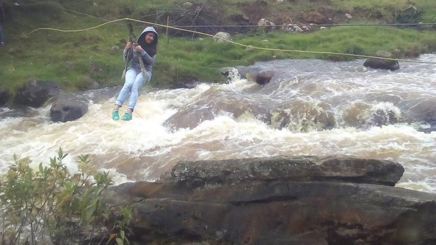 Persona atravesando un río de corriente fuerte mediante una cuerda, rodeada de rocas y vegetación en un entorno rural.