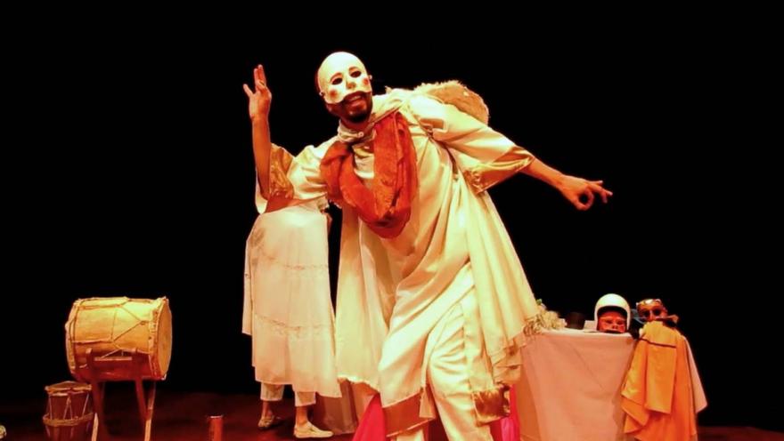 Un artista con máscara blanca y vestuario tradicional protagoniza una escena teatral llena de expresión, música y simbolismo, acompañado de tambores y otros elementos escénicos.