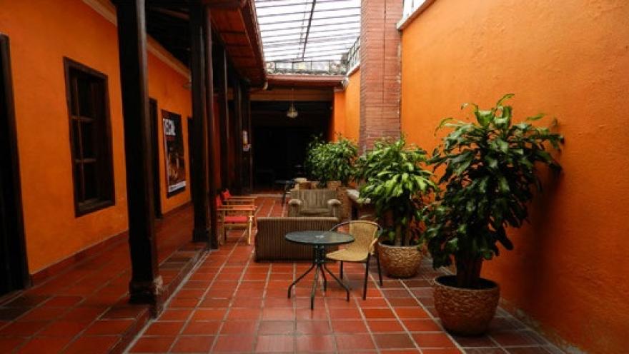 Patio interior de arquitectura colonial, con paredes naranjas, corredores de madera, plantas ornamentales y mesas que crean un ambiente acogedor y tranquilo.