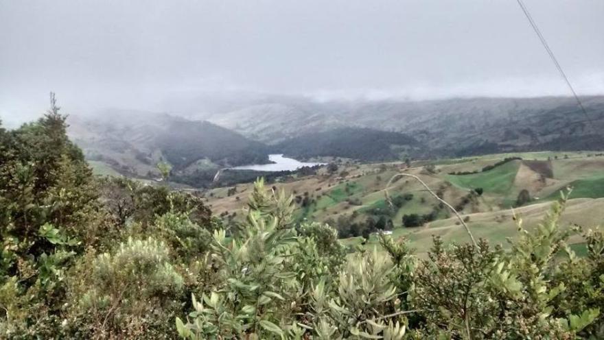 Vista panorámica de montañas verdes, vegetación nativa y terrenos rurales, con una laguna al fondo bajo un cielo nublado.