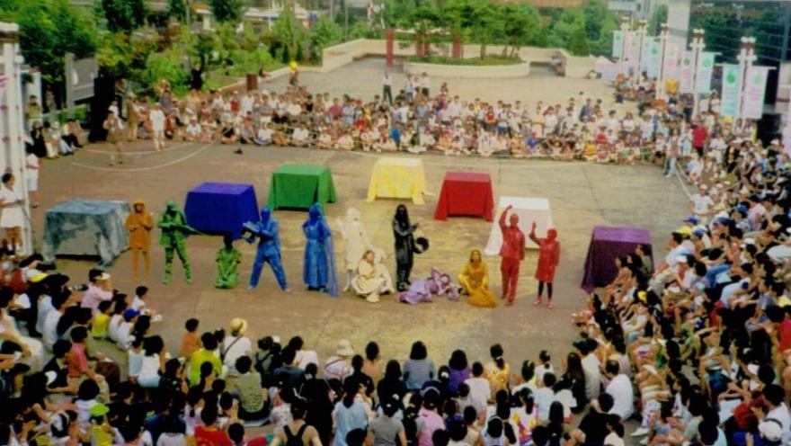  Público reunido en una plaza para disfrutar de una presentación de teatro callejero, con artistas vestidos de colores y una puesta en escena al aire libre.