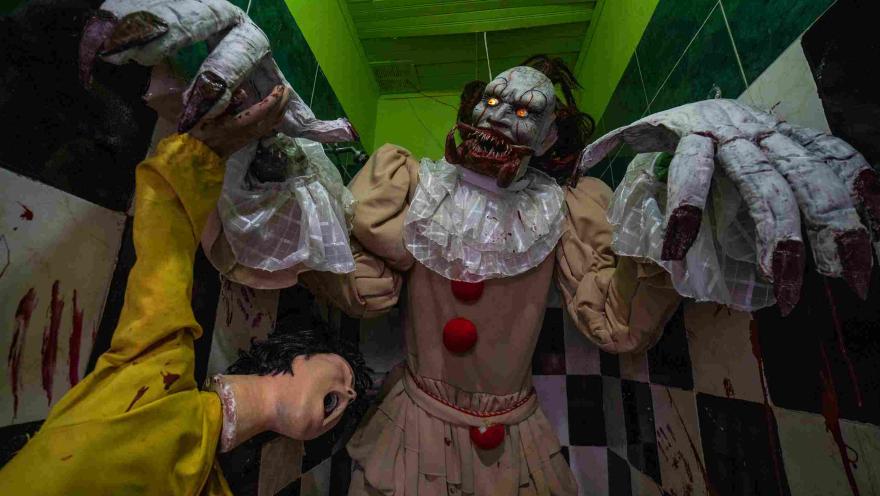 Escenario de terror del Museo de la Selfie en Bogotá, con una figura de payaso siniestro en el centro, acompañada por criaturas de aspecto monstruoso con grandes manos y rostros aterradores. El espacio está ambientado con paredes verdes, un piso de cuadros blancos y negros y elementos que recrean una escena de horror.