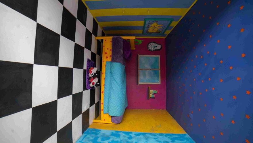  Escenario de ilusión óptica del Museo de la Selfie en Bogotá, con una habitación decorada en colores azul, amarillo y rojo. Una cama parece estar dispuesta en una posición inclinada, mientras las paredes presentan patrones de estrellas y cuadros en blanco y negro que generan un efecto visual sorprendente.