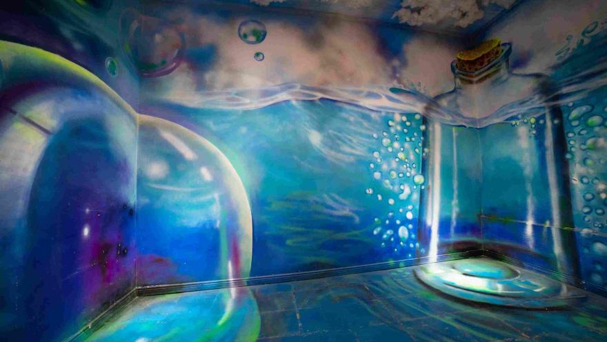 Escenario inmersivo del Museo de la Selfie en Bogotá que recrea un mundo submarino, con paredes y piso decorados con imágenes de agua, burbujas, peces y objetos flotantes. La iluminación en tonos azules y verdes crea la sensación de estar bajo el agua.