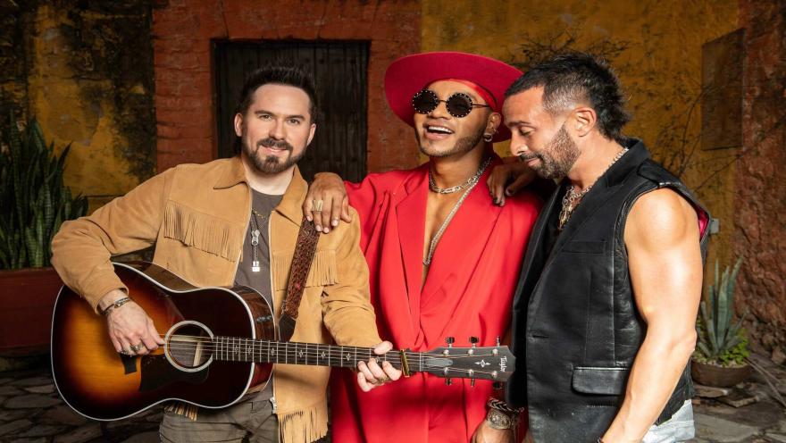 Los tres integrantes del grupo Camila posan juntos; uno de ellos sostiene una guitarra acústica en un patio.