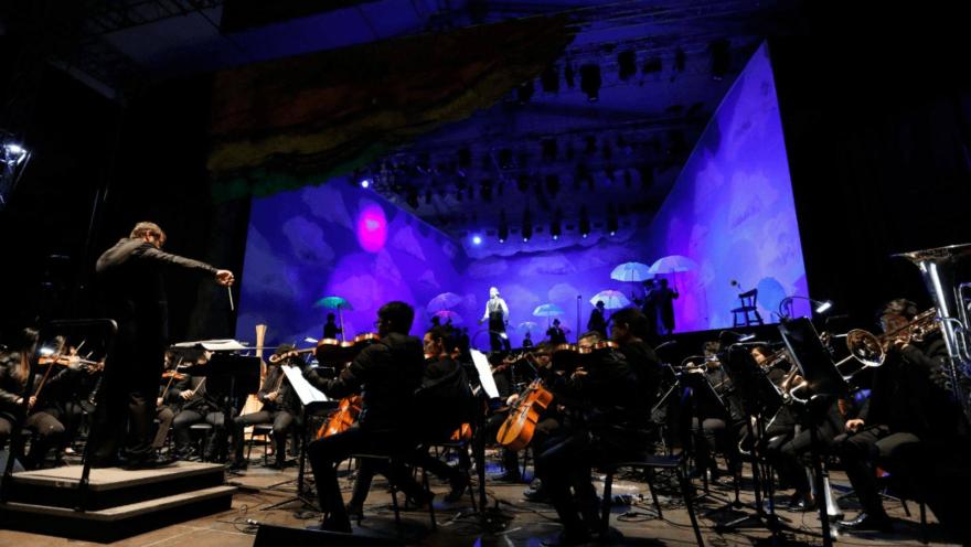 Una gran orquesta filarmónica interpreta una obra en escenario iluminado con tonos azules durante una función.