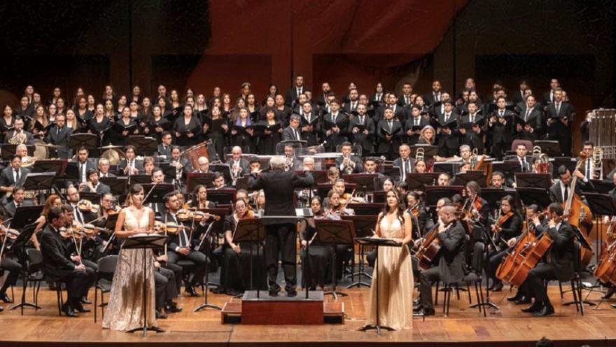 Gran montaje orquestal y coral sobre el escenario con solistas al frente y director en el podio.