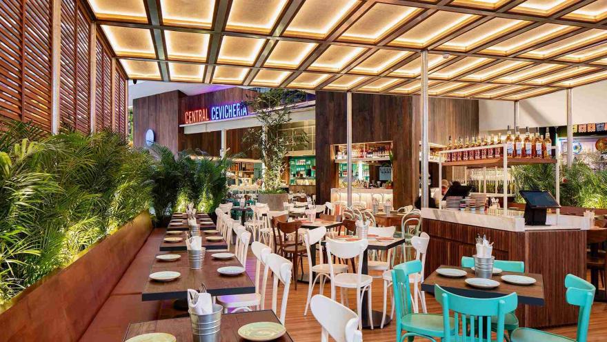Amplio salón del restaurante con mesas de madera, sillas blancas y turquesas, plantas verdes y techo iluminado.