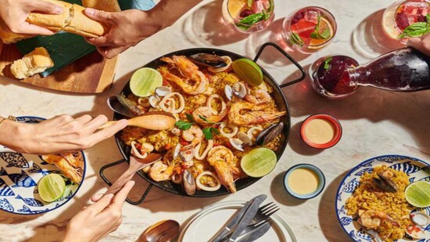 Vista superior de una paella marinera con camarones, mejillones y calamares rodeada de comensales y bebidas.