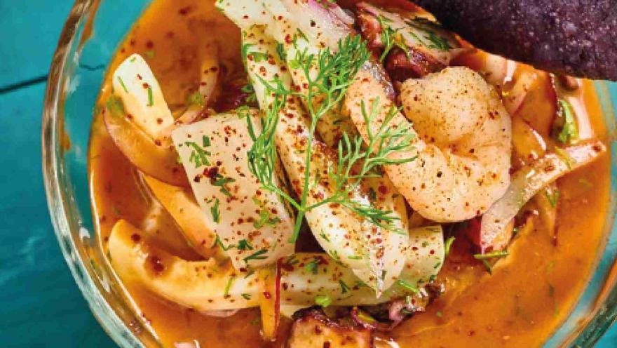 Primer plano de ceviche servido en tazón de vidrio con camarones, pescados, pulpo, especias y hierbas frescas.