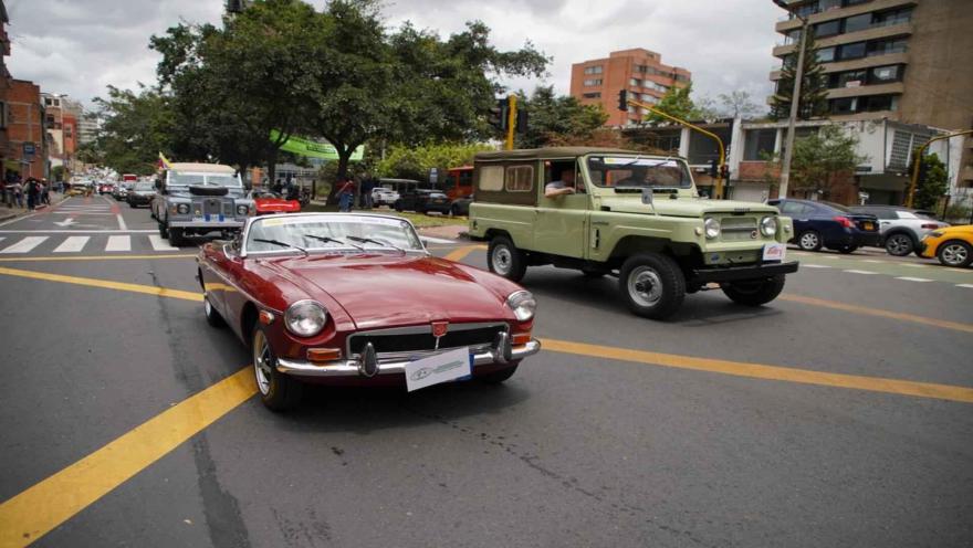 Un descapotable clásico rojo y un campero antiguo verde transitan en un desfile por una calle de Bogotá.