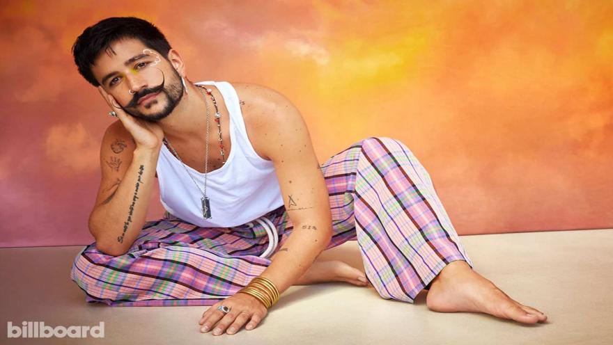 El cantante Camilo, con su característico bigote y ropa colorida, se apoya en su mano sentado en el suelo.