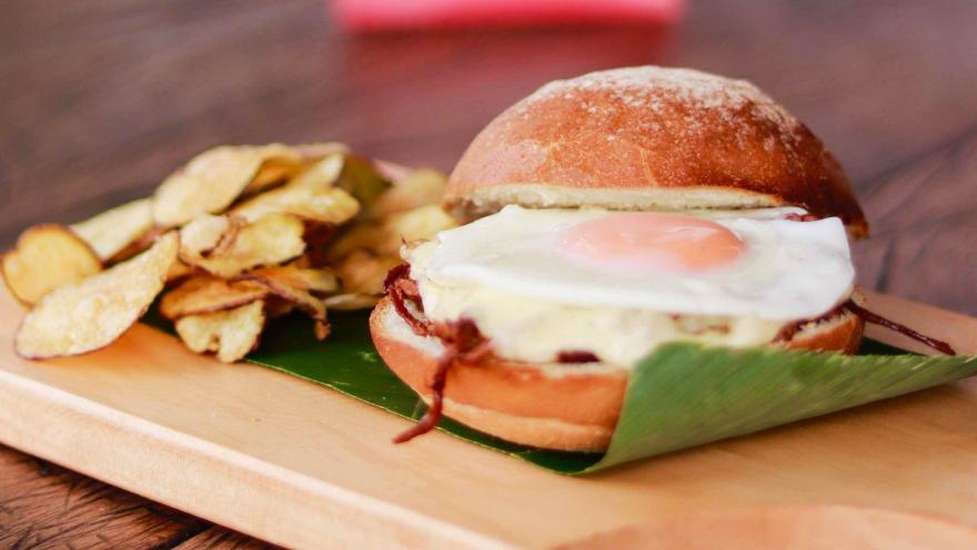 Hamburguesa con huevo frito, queso y tocineta, acompañada de chips de papa sobre una tabla de madera.