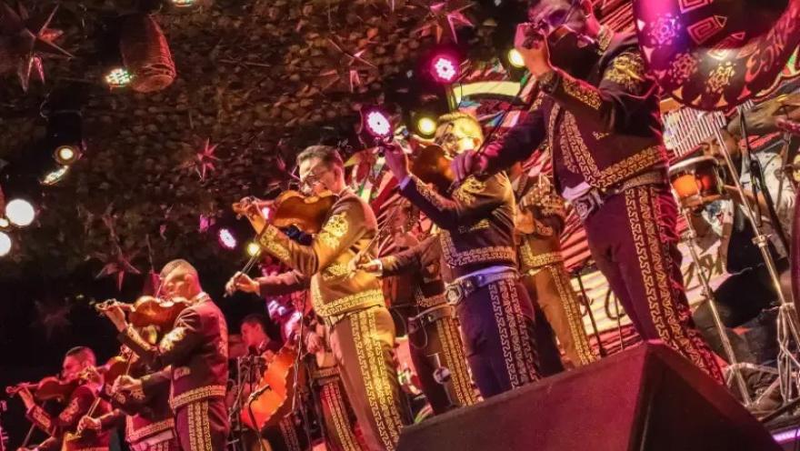 Grupo de mariachis interpretando música en vivo sobre un escenario, con iluminación colorida y ambiente festivo.