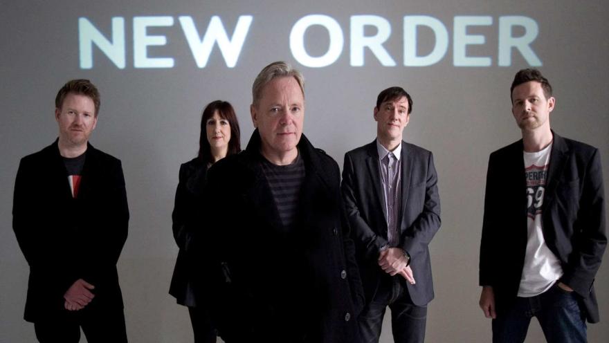 Los integrantes de la banda de rock New Order posan de pie con atuendos oscuros frente a una pantalla con su nombre.