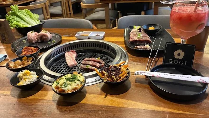 Mesa de barbacoa coreana con parrilla central, cortes de carne, acompañamientos, salsas y una bebida refrescante.