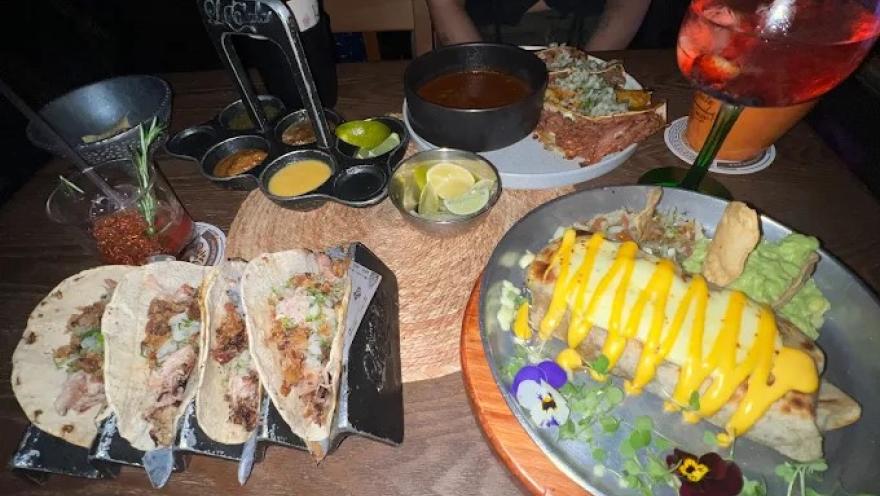 Mesa con tacos, platos de comida mexicana, salsas, guacamole y bebidas, en un ambiente de celebración.