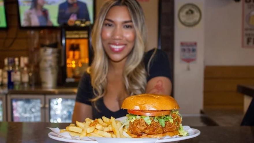 Mujer joven muestra un plato de Hamburguesa acompañada de papas fritas, servida en mesa de Hooters.